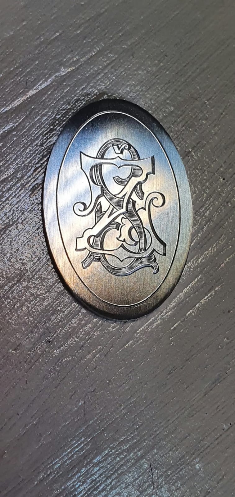 Ovalna jeklena ploščica z ročno graviranim prepletenim monogramom.